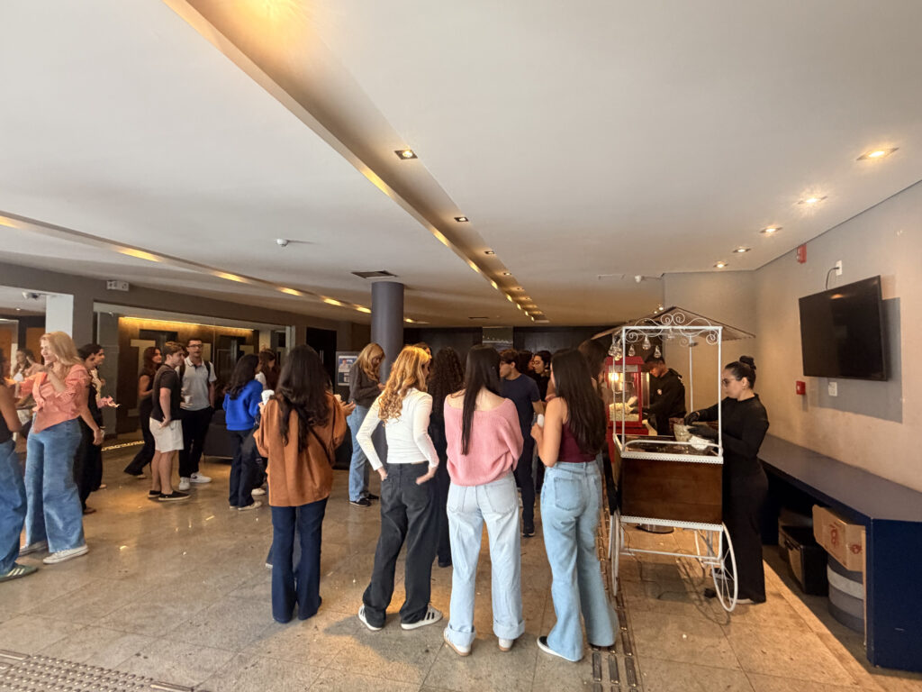Pessoas em fila em um foyer com luzes embutidas e carrinho de doces, algumas de costas conversando enquanto aguardam atendimento em um evento interno.