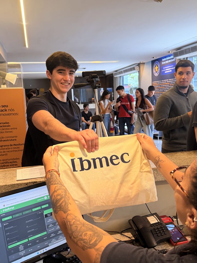 Jovem exibem uma ecobag com o logo “Ibmec” na recepção da instituição ao lado de computadores e pessoas ao fundo.