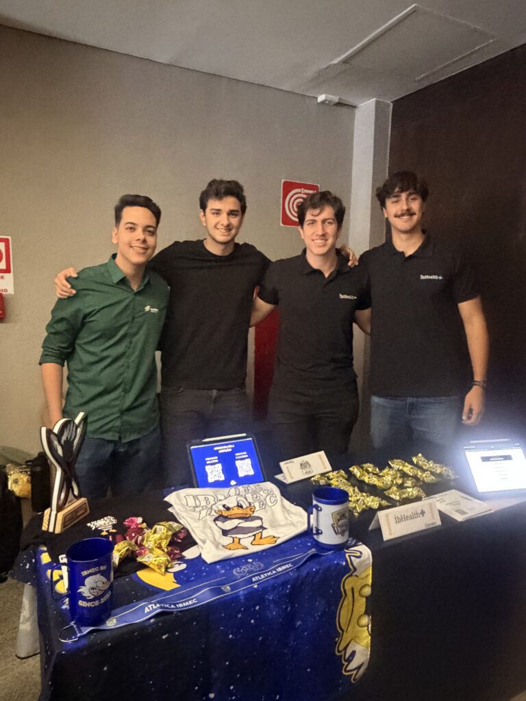 Equipe de quatro homens em evento corporativo do Ibmec, posando atrás de uma mesa com brindes, chocolates embrulhados e canecas; ao fundo há cartazes e iluminação do estande.