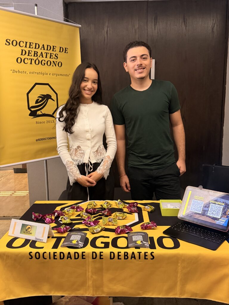 Foto de dois estudantes em evento com banner “SOCIEDADE DE DEBATES OCTÓGONO” e mesa amarela com doces e notebook, em um cenário de conferência.