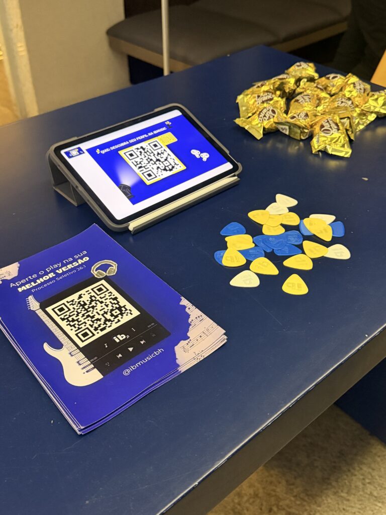 Mesa com material promocional de uma campanha do IbMusic: QR codes em cartões azuis, tablet exibindo código e moedas de chocolate douradas e amarelas ao lado de um papel com instruções.