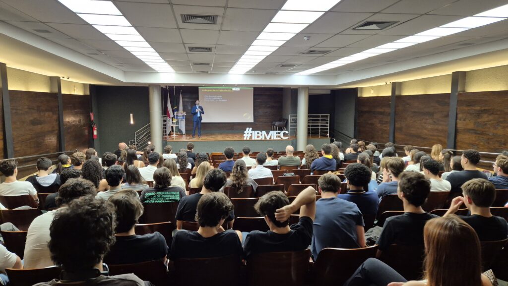 Público em uma palestra em auditório lotado, com professor em um palco e banner “#IBMEC” ao fundo, durante aula magna para calouros.