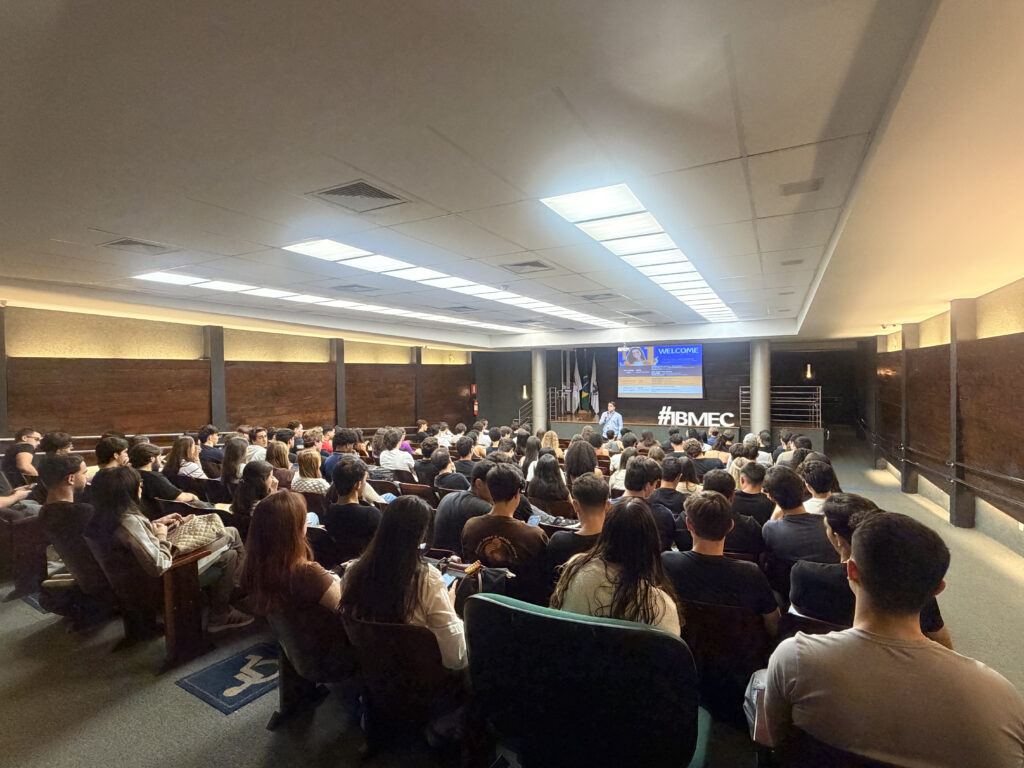 Público sentado em auditório durante palestra em evento com palco e telão ao fundo, iluminação do teto e banner com a hashtag #IBMEC na conferência.