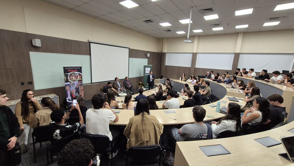 Sala de aula ou auditório durante uma reunião: participantes sentados ao redor de uma mesa grande com computadores e anotações, apresentador em pé diante de um telão com projetor