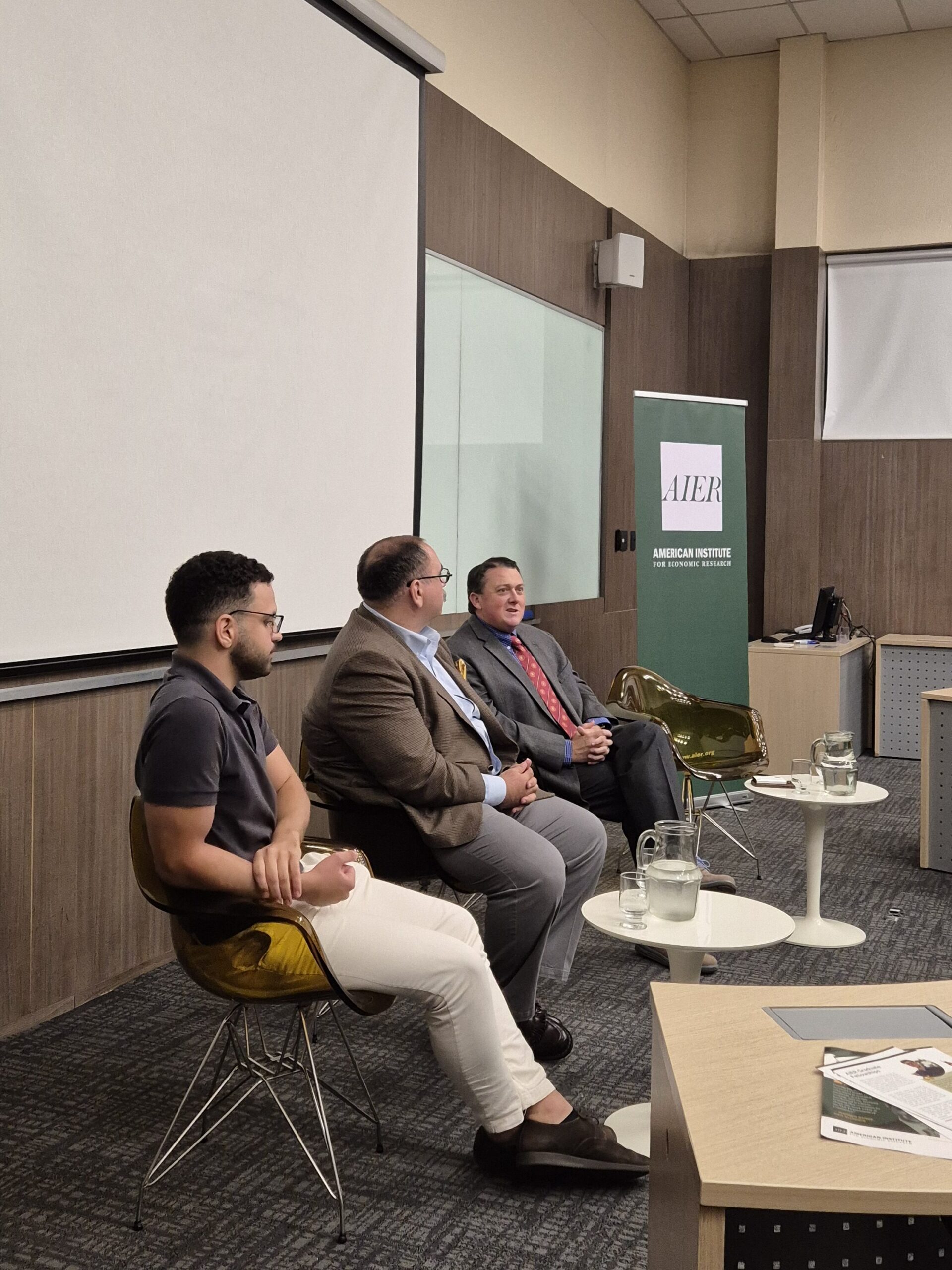 Palestra em auditório com três participantes sentados em cadeiras e apresentador em sala de conferências, banner do AIER ao fundo.