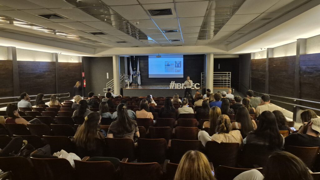 Sala de auditório lotada durante palestra, com público sentado e telão central exibindo slide; no palco há estrutura de apresentação e faixa #IBMEC.