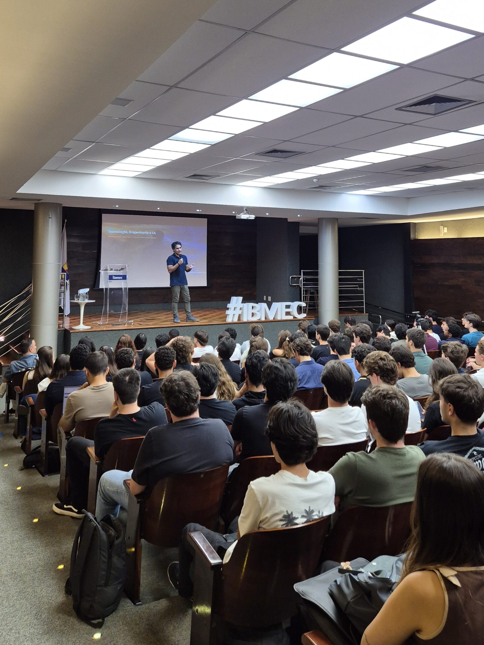 Palestra em auditório com plateia sentada, telão ao fundo e apresentador em palco durante o evento #IBMEC, com teto em painéis de iluminação