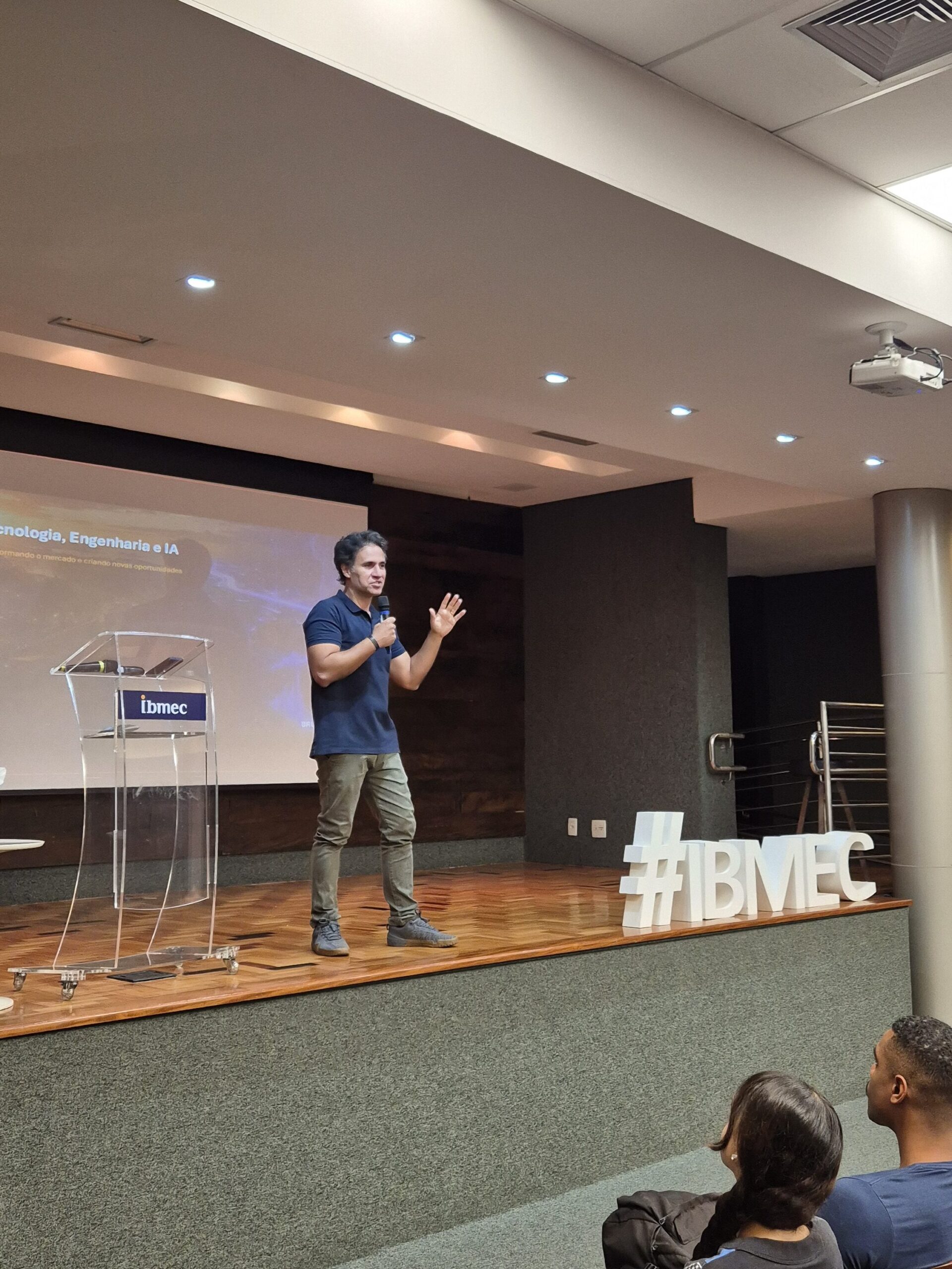 Palestra no auditório do Ibmec: palestrante segura microfone no palco e apresenta para o público, com telão ao fundo e letreiro #IBMEC no palco.