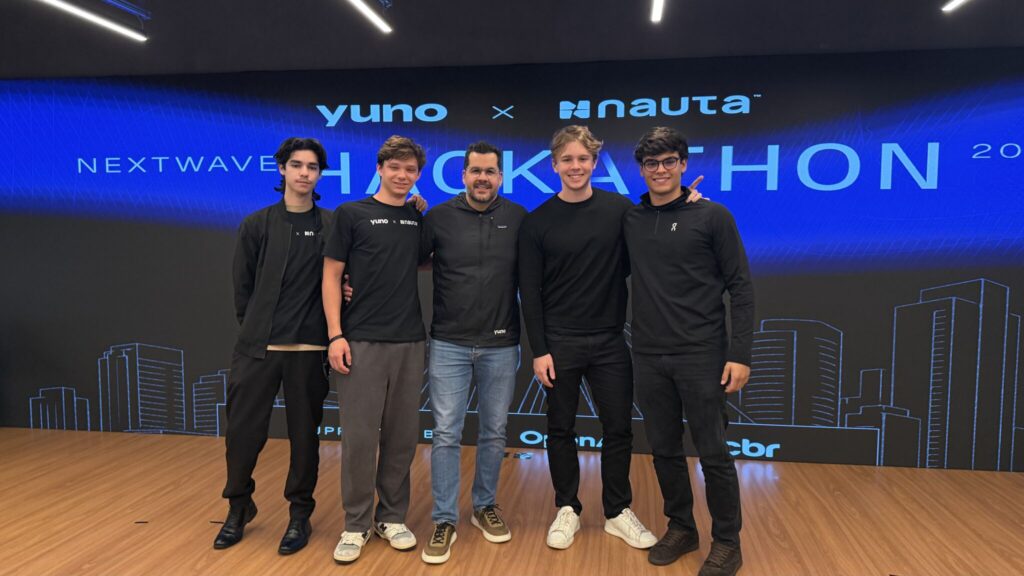 Equipe Yuno e Nauta e alunos Ibmec BH posando em palco durante evento, com telão azul ao fundo e skyline estilizado; cinco pessoas vestidas de preto e jeans em frente à marca Yuno X Nauta.
