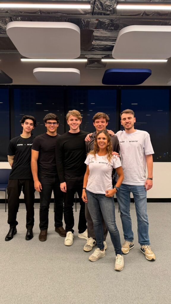 Equipe sorridente posa em um ambiente de escritório com iluminação no teto e displays azuis ao fundo, usando camisetas com logo da empresa.