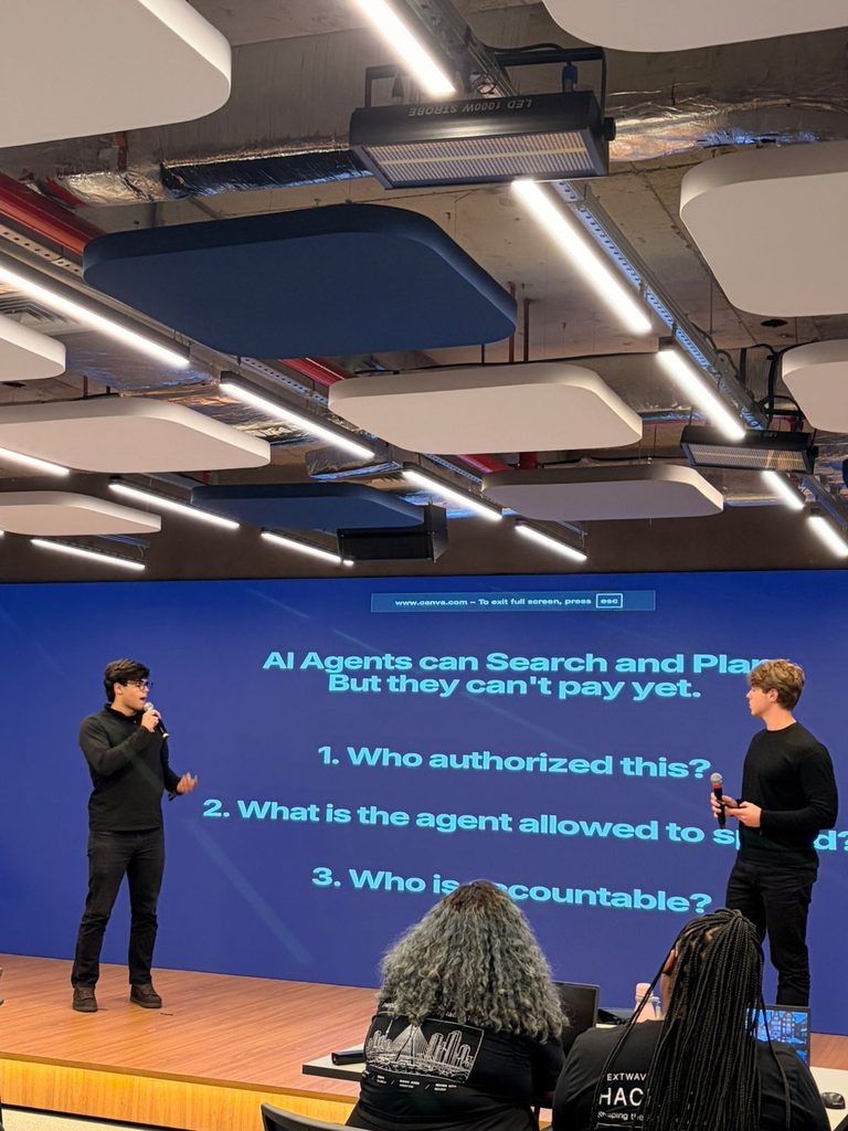 Palestra em auditório com dois alunos apresentando de pé no palco, telão azul exibindo texto em inglês sobre agentes de IA e perguntas enumeradas, iluminação do teto e público em primeiro plano.