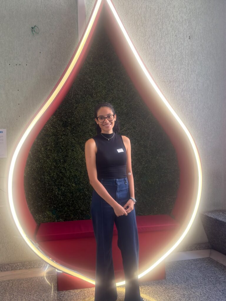 Aluna em pé sorrindo em frente a um grande recorte em formato de gota com luzes de LED, com fundo de painel verde e paredes claras no ambiente interno.