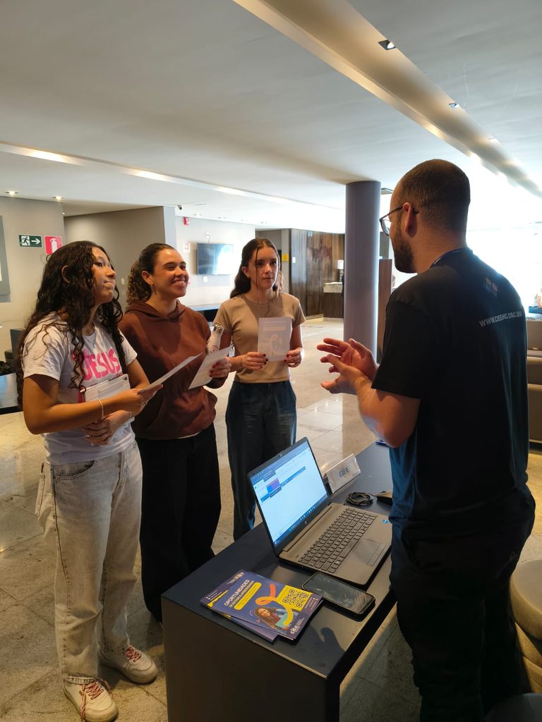Equipe apresentando projeto em evento, com pessoas segurando folhetos e um monitor exibindo conteúdo em um balcão, em ambiente interno com iluminação do teto.