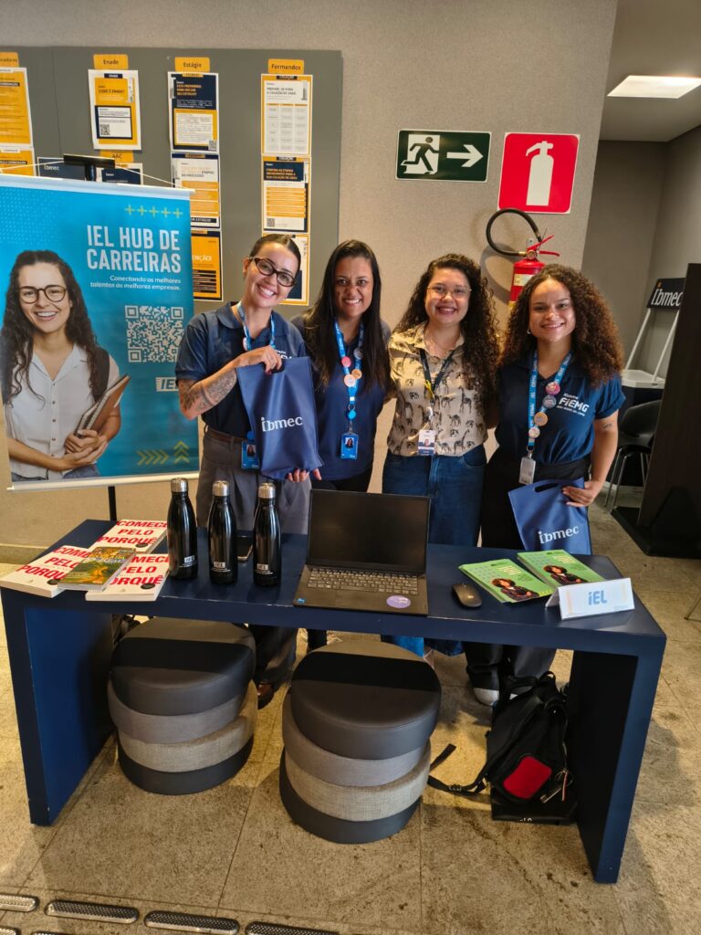 Equipe sorridente no estande do IEL Hub de Carreiras durante evento, com banner informativo ao fundo e mesa com garrafas e materiais de divulgação.