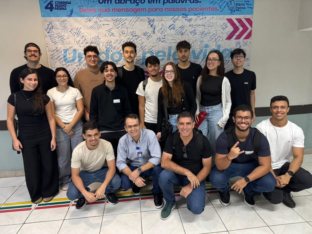 Foto de grupo de participantes de evento corporativo, com cerca de dez pessoas em pé e cinco sentadas no chão, sorrindo, em frente a um mural com fundo azul em hospital.