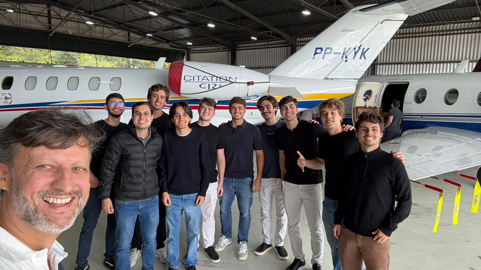 Grupo de homens sorrindo em frente a aeronaves no hangar, com um grande painel do avião ao fundo e equipamentos de aviação ao lado.