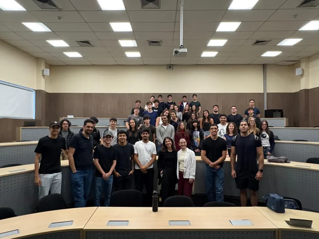 Grupo de pessoas em uma sala de aula ou auditório, posando para foto em frente a uma mesa grande, com iluminação de teto e projetor no ambiente.