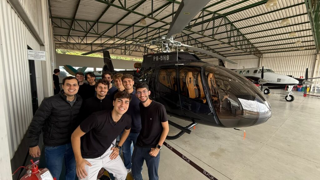 Grupo de jovens em frente a um helicóptero na área interna de um hangar, com o rotor e o casco visíveis ao fundo e a equipe sorrindo para a foto.