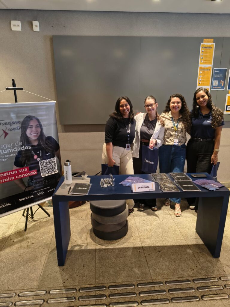 Foto de quatro pessoas sorrindo e posando atrás de uma mesa com materiais e brindes em um evento, com banners e cards informativos ao fundo.