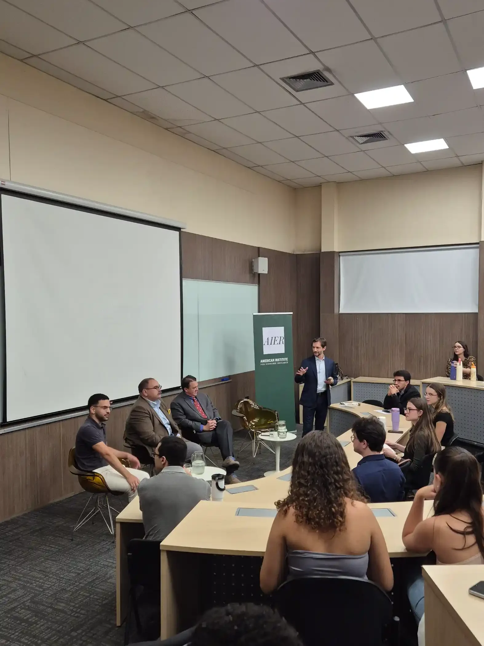 Reunião em sala de treinamento: palestrante em pé apresenta conteúdo para um grupo sentado em volta de mesas, com telões de projeção e mesas organizadas.