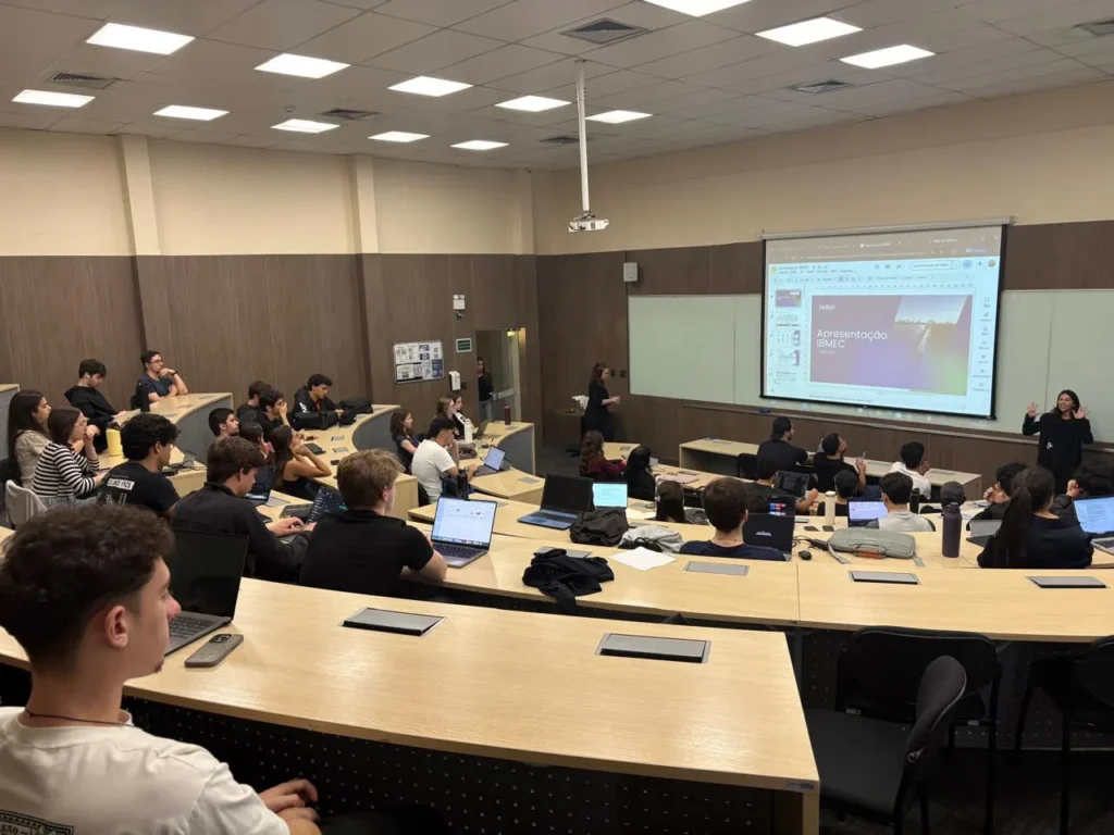 Sala de aula com turma de estudantes reunidos em carteiras, alguns usando notebooks, enquanto um professor apresenta uma apresentação no telão