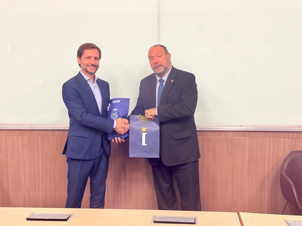 No El Salvador Day, dois homens, Adriano Gianturco e Luís Aparicio, em trajes sociais, fazem a saudação de aperto de mãos durante cerimônia em ambiente corporativo, com mesa e quadro ao fundo.