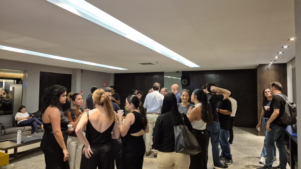 Grupo de pessoas conversando em um evento em um ambiente interno, com cadeiras ao fundo e iluminação de teto em destaque, clima social e descontraído.
