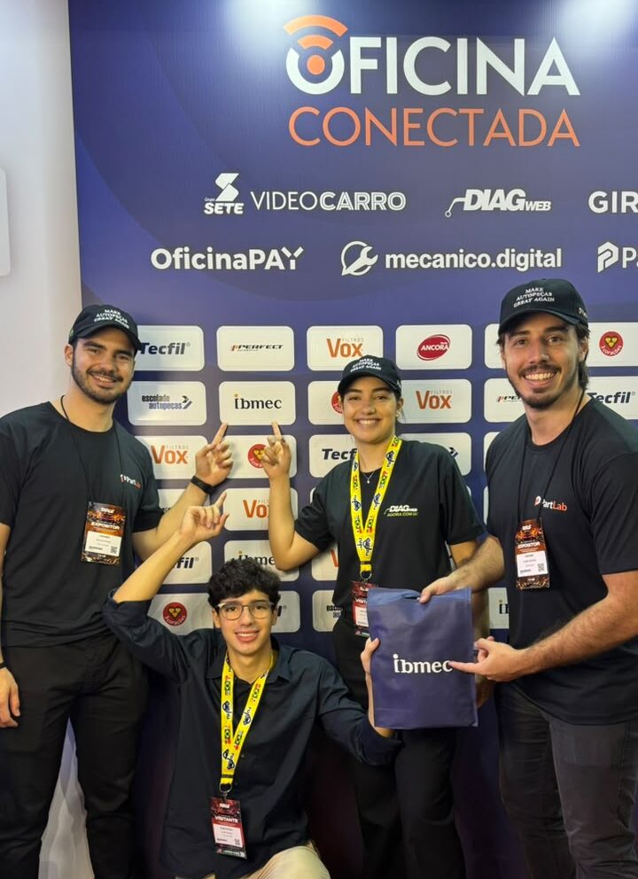 Equipe de participantes em evento de oficina, com três homens em pé e uma pessoa agachada em frente a um painel azul exibindo medalhas e brindes.
