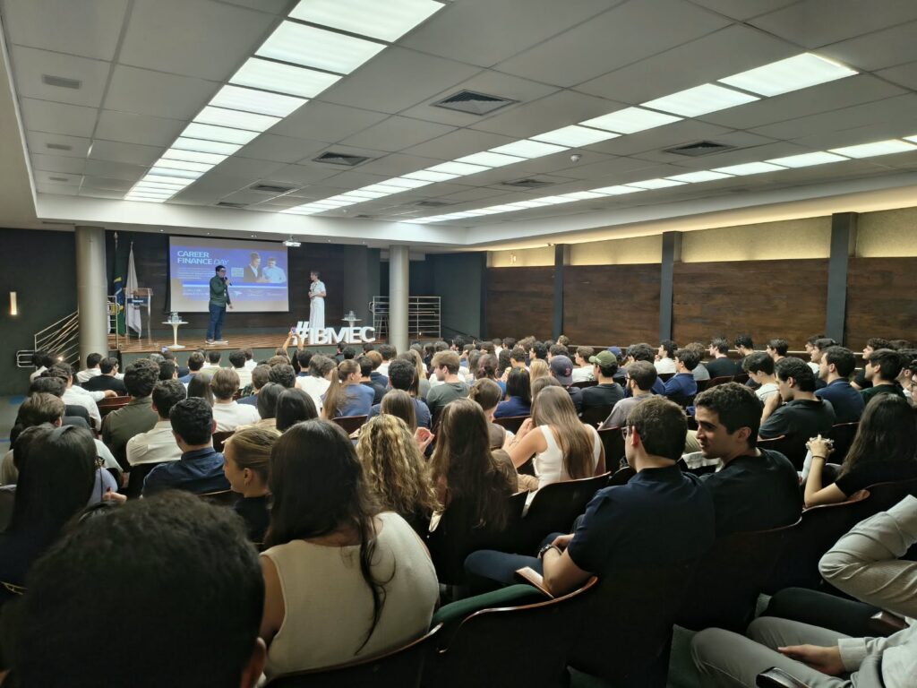 Público em um auditório durante palestra em evento corporativo, com apresentadores em palco e telão ao fundo mostrando informações do encontro. Assentos lotados.