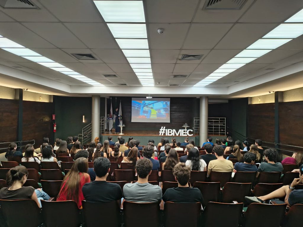 Palestra em auditório lotado com telão exibindo apresentação e banner #IBMEC ao fundo, durante evento acadêmico com público sentado em cadeiras.