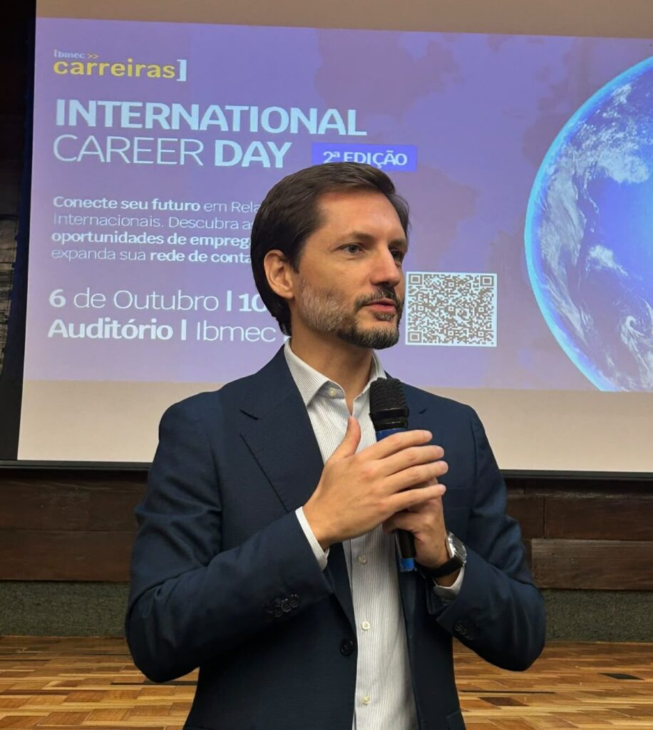 Professor Adriano Gianturco palestrando em evento corporativo com microfone na mão, em frente a um telão com o banner “INTERNATIONAL CAREER DAY” e QR code.