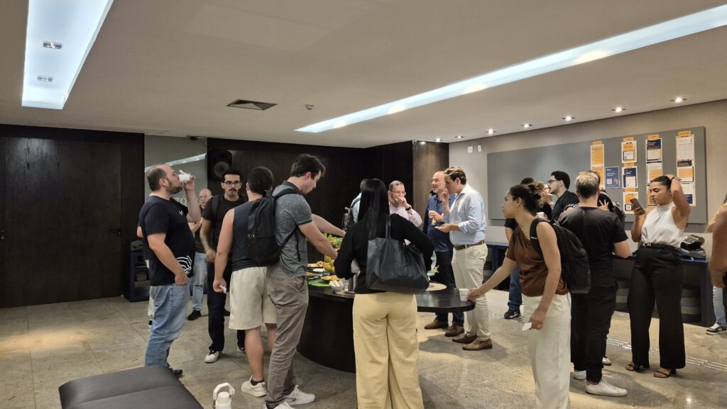 Foto de pessoas reunidas em um evento em uma sala de recepção, conversando em pé perto de uma mesa com buffet, com cartazes informativos ao fundo e iluminação embutida no teto.